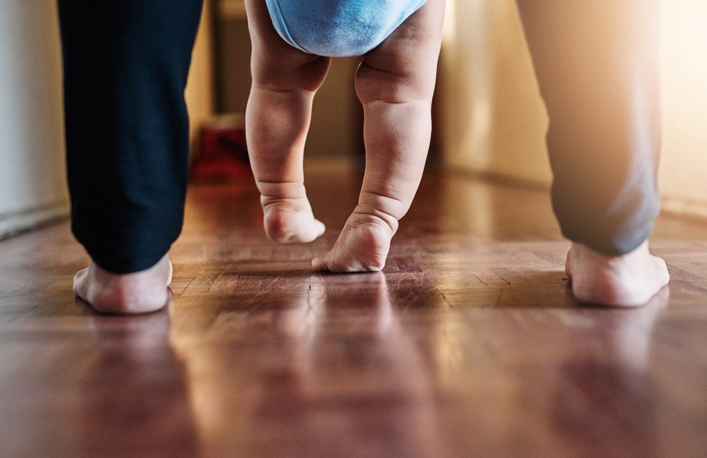 A baby learning to walk with support from a parent; only their feet are shown.